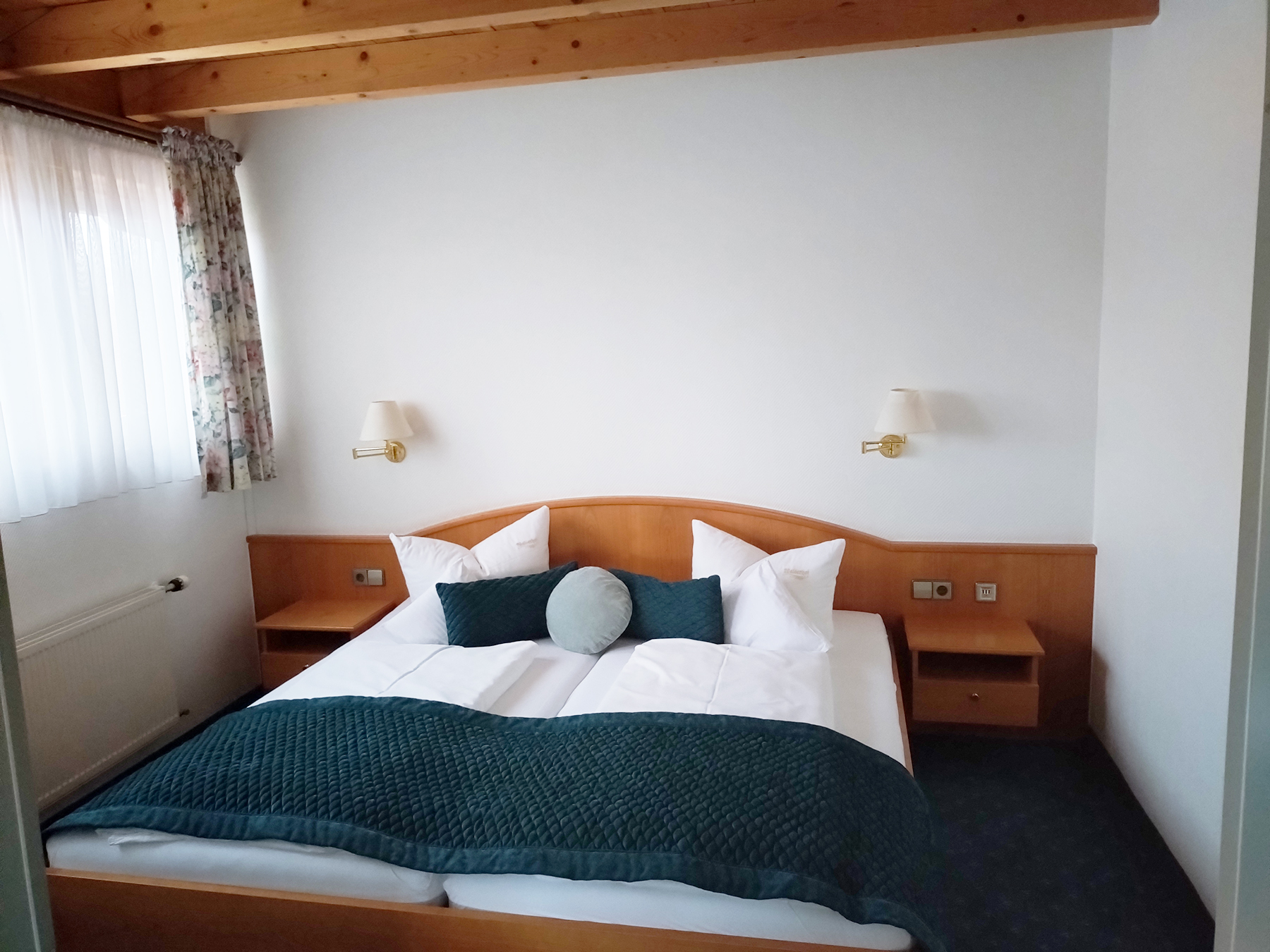 Apartment-Schlafraum mit Doppelbett, weißen Bettlaken, Holznachttischen und Fenster mit Gardine